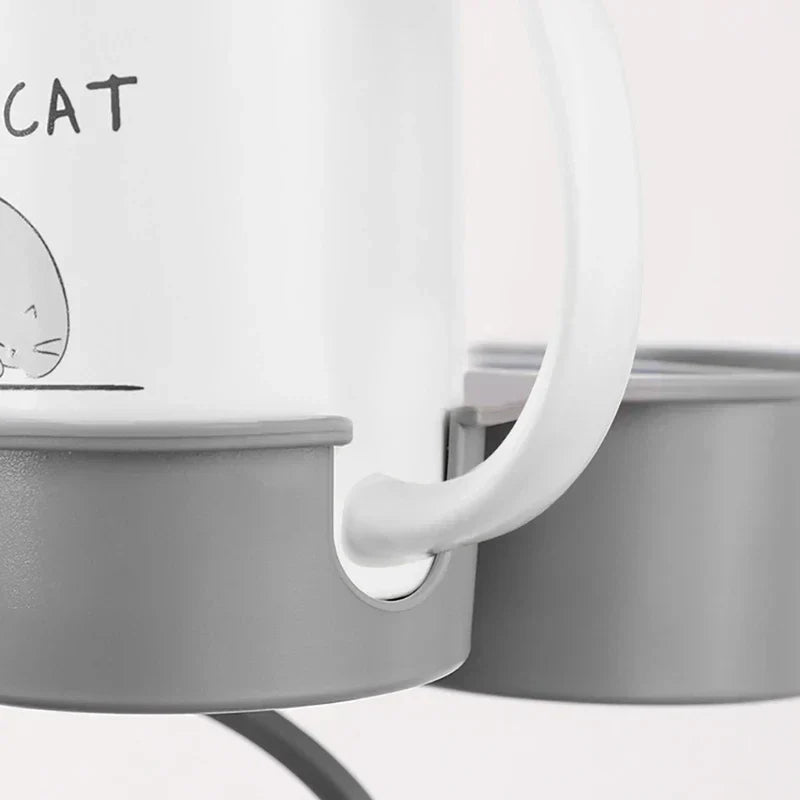 Soporte con pinza para bebidas de mesa - Primer plano de una taza blanca con un gráfico parcial de gato y texto, descansando en un soporte de plástico gris junto a un contenedor a juego