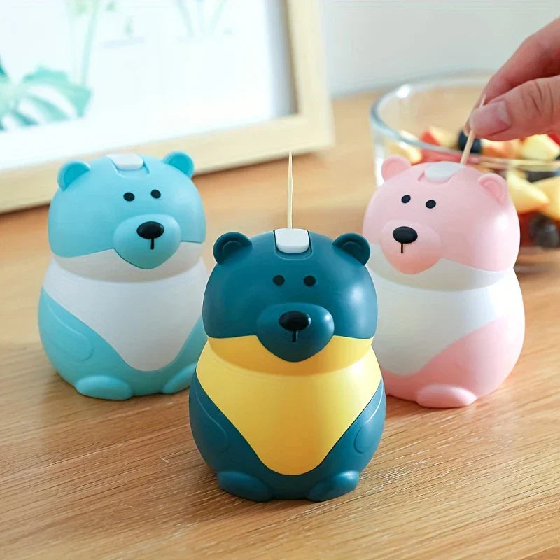 Sujetapalillos con forma de osito de peluche: tres sujetapalillos multicolores con forma de oso sobre una mesa de madera, con una mano sacando un palillo del sujetapalillos rosa y un frutero al fondo