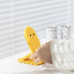 Folding Phone Stand - Hand adjusting the angle of the yellow chick design stand placed on a sunlit desk