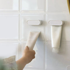 Toothpaste Tube Squeezer - Two white wall clips mounted on a white tiled wall; one holds a squeeze tube, a hand inserts a second tube into the empty clip