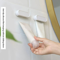 Toothpaste Tube Squeezer - White wall-mounted clip holders, one holding a white tube, hand inserting a second tube into the empty clip.