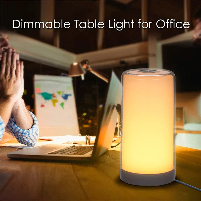 Warm lit led desk lamp on an office desk with a laptop and a person in the background