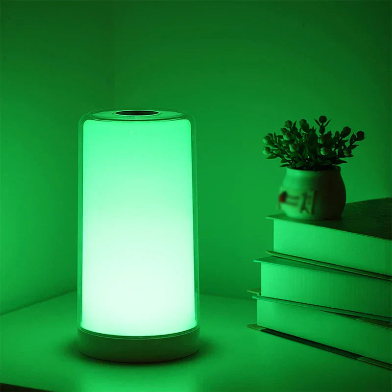 Green glowing led desk lamp sits on a table beside stacked books with a small potted plant on top.