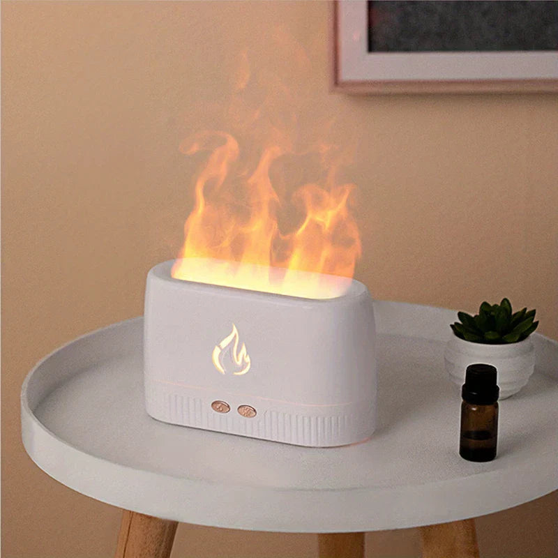 Ultrasonic Humidifier - White matte flame effect aroma diffuser sits on white round side table, emitting simulated orange flame mist, with essential oil bottle and small succulent nearby.