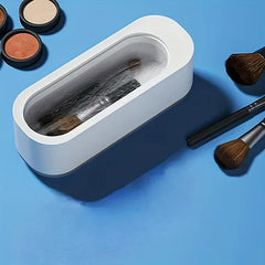 Máquina limpiadora ultrasónica: recipiente rectangular blanco de apertura superior que contiene brochas de maquillaje en líquido ondulante, con artículos de maquillaje adicionales sobre superficie azul