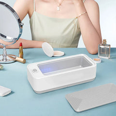 Ultrasonic Glasses Cleaner Machine - Open white ultrasonic cleaner on a light blue vanity table, its ridged grey lid placed beside it, with cosmetics, a mirror, and a partially visible woman in the background.