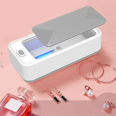 Ultrasonic Glasses Cleaner Machine - White and gray ultrasonic cleaning machine with lid propped open, blue indicator lights lit, beside jewelry and perfume bottle on pink surface