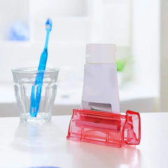 Wall Mount Toothpaste Dispenser - Translucent red toothpaste tube squeezer holding a partial white toothpaste tube, with a glass and blue toothbrush in soft background