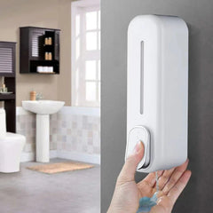 A thumb presses the button of a white wall-mounted soap shampoo dispenser, dispensing liquid into a palm, with a bathroom in the background.