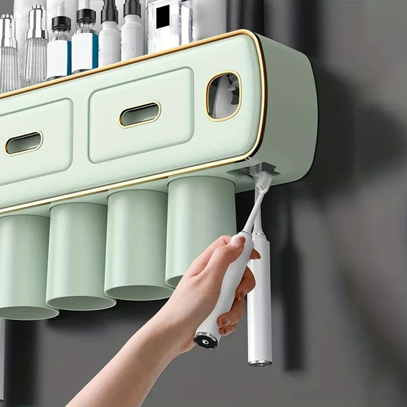 Hand uses an electric toothbrush with a mint green, gold-accented wall mount toothbrush holder toothpaste dispenser set holding inverted cups and toiletries on top