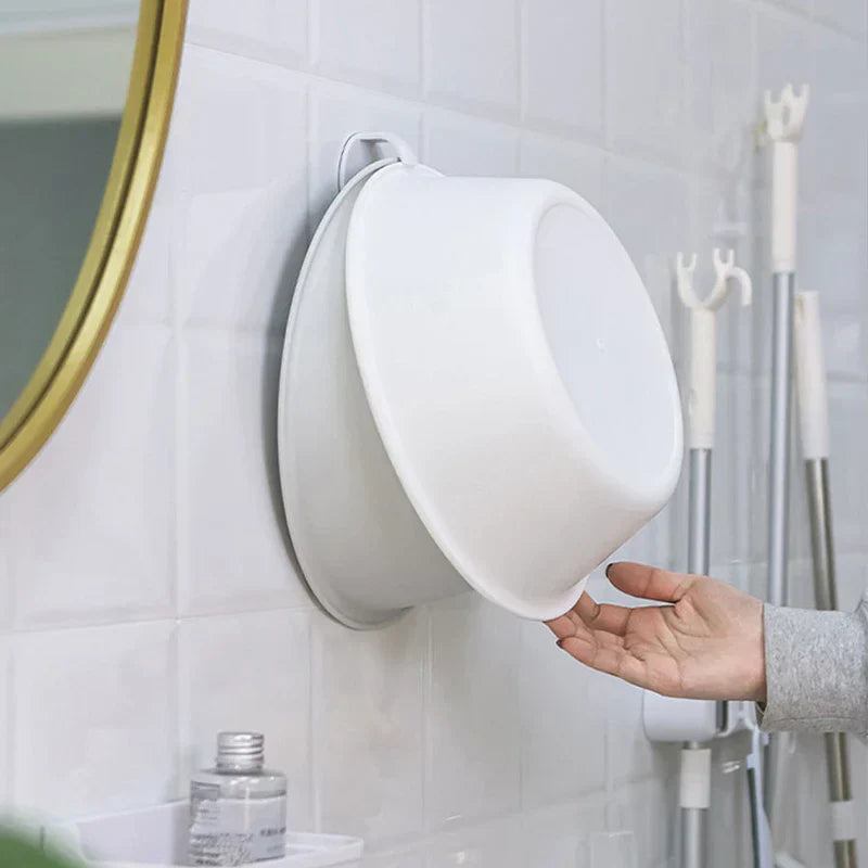 A white wash basin storage rack mounted on a white tile bathroom wall holds a white plastic basin, with a hand reaching for the basin.