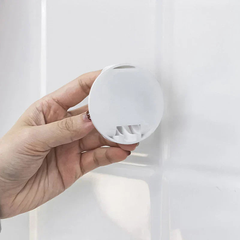 A hand holds the back of a small white circular adhesive hook; the wash basin storage rack is not visibly present.