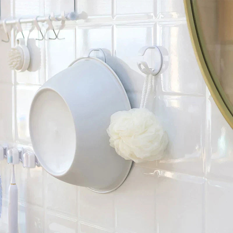 A light gray basin hangs on a white wall hook above a tiled bathroom wall, with a bath puff hanging on an adjacent hook; a wash basin storage rack is on the tile wall holding the basin.