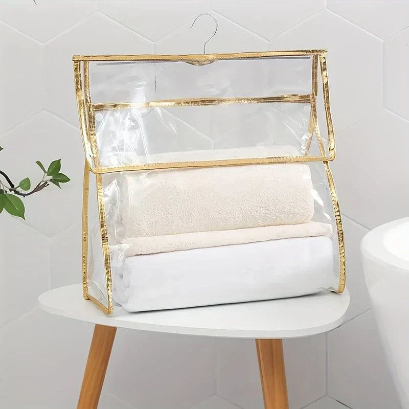 Shower Organizer - Clear hanging storage bag with gold trim holding folded white and cream towels, resting on a small white side table