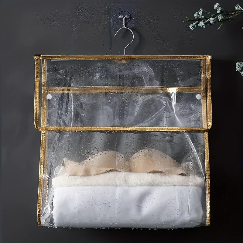 Shower Organizer - Clear hanging storage bag with gold trim holding folded linens and a bra, hung on a dark wet wall with visible water droplets on the bag surface