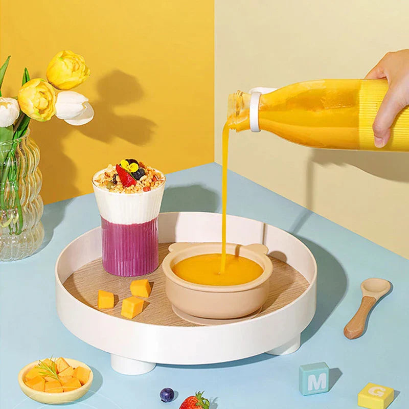 Drinking Glass - Hand pouring yellow liquid from a yellow ribbed bottle into a small bowl, tray with layered cup, mango cubes, tulip vase on blue table