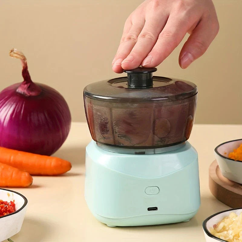 Picadora de alimentos eléctrica - una mano presiona el botón superior de la picadora sobre una encimera, con zanahorias y cebolla colocadas a su lado.