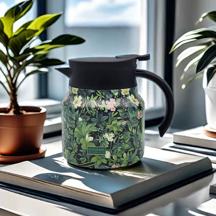 Vintage Floral Tea Thermos - Insulated pitcher with dark green leafy floral print, black lid and curved handle, placed on a book near potted plants by a window