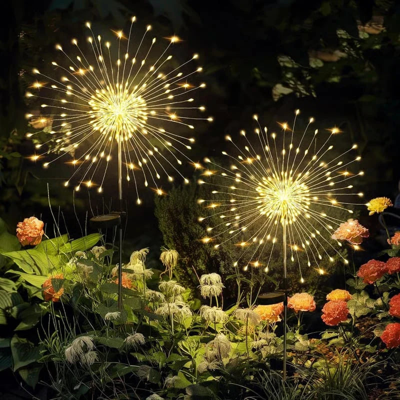 feenlichter - Zwei helle solarbetriebene Garten-Feenlichter stehen nachts zwischen Gartenblumen und Laub und strahlen ein warmes goldenes Leuchten aus.