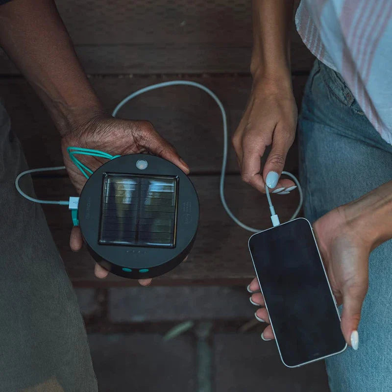 Two people use a solar camping lantern to charge a smartphone via a USB cable outdoors