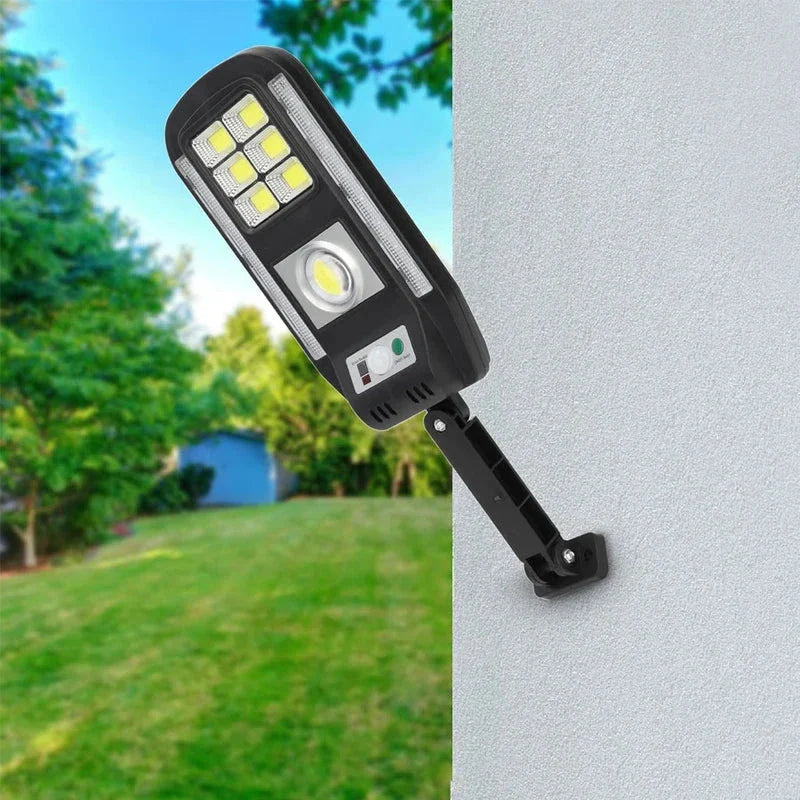 Lámpara de seguridad con sensor de movimiento alimentada por energía solar montada en una pared exterior gris, con un jardín de césped y árboles desenfocados al fondo.