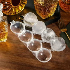 Clear silicone ice tray tupperware with four empty and four frozen spherical ice molds sits on a wooden table near glass decanters.