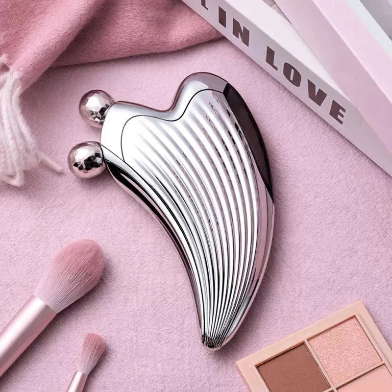 Beauty Device - Shiny silver heart-shaped facial massager resting on pink fabric, with makeup brushes and an eyeshadow palette nearby