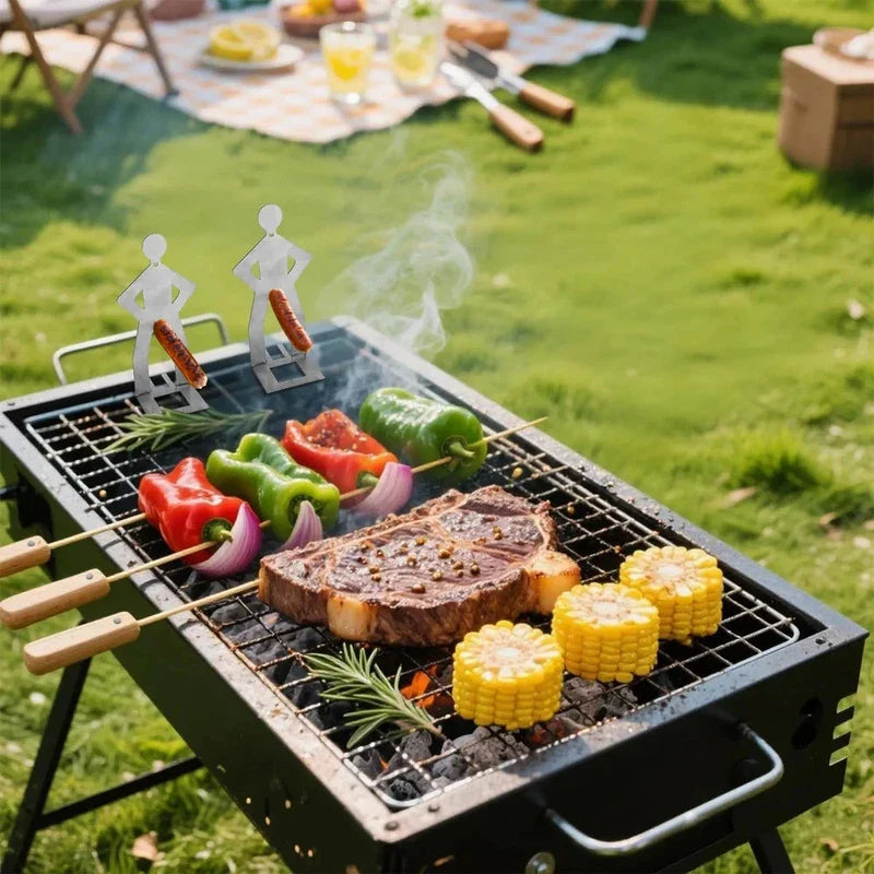 Funny Sausage Grill - Two stainless steel human-shaped sausage holders on a portable charcoal grill, with steak, vegetables, and corn cooking beside them