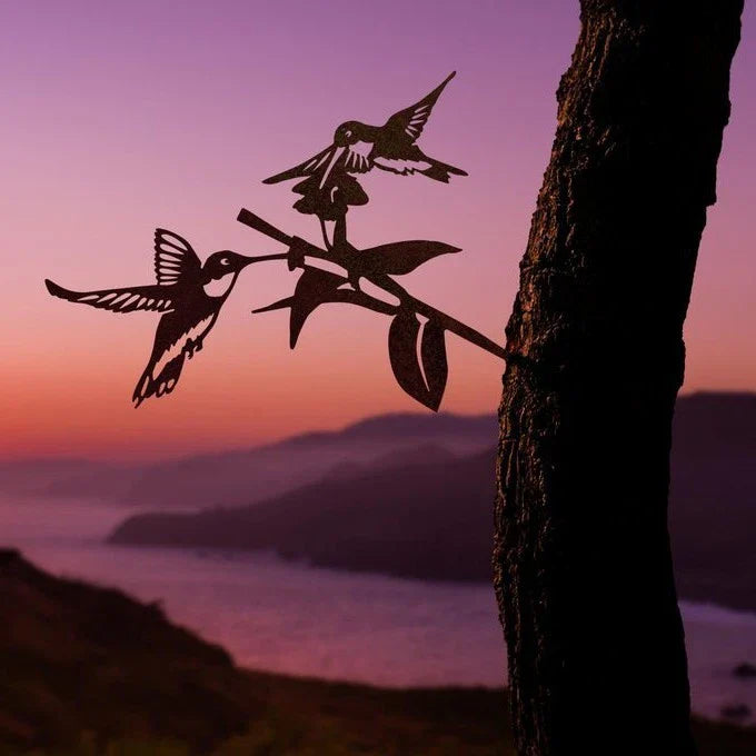 Silhouette of two hummingbirds on a flowering branch mounted to a tree, with a purple sunset and water in the background.