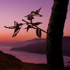 Silhouette of two hummingbirds on a flowering branch mounted to a tree, with a purple sunset and water in the background.