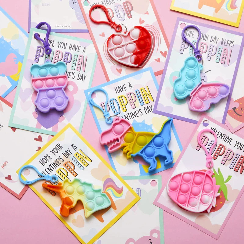 Six assorted valentines day cards with pop fidget keychains laid flat on a pink background.