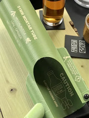 Close-up of green 51mm mortar tube themed bottle opener with printed labeling and caution graphic on its opening.