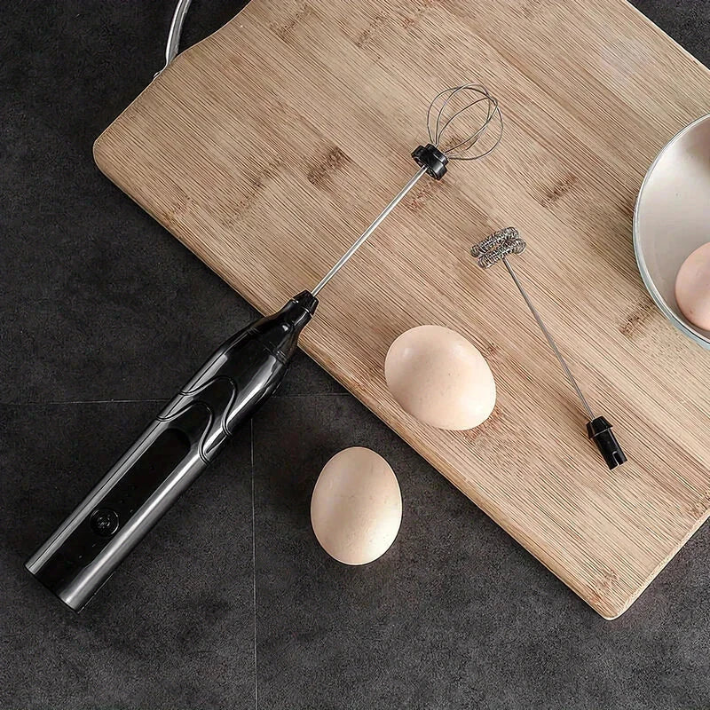 Electric Hand Mixer - Black handheld electric whisk with two interchangeable heads, resting on a cutting board beside two eggs and a partial bowl of egg.