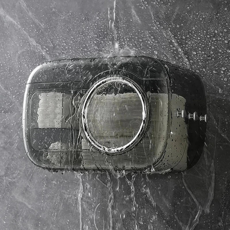 Cotton Pad Dispenser Holder - Closed transparent gray wall-mounted dispenser with water streaming over it, droplets visible, mounted on a wet gray tiled surface with white rolls inside.