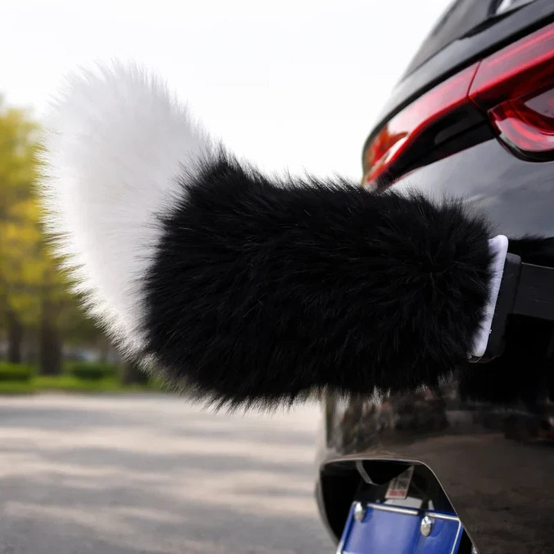 Black and white fluffy car moving tail attached to the rear of a black car near the taillight, with a blurred outdoor background.