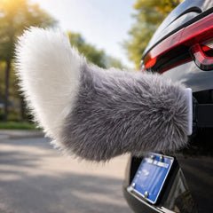 Gray and white fluffy car moving tail attached to the rear of a black car near the taillight, with a sunny blurred outdoor background.