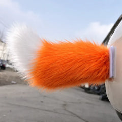 A car moving tail with orange and white faux fur attaches to a white car outdoors near a paved area.