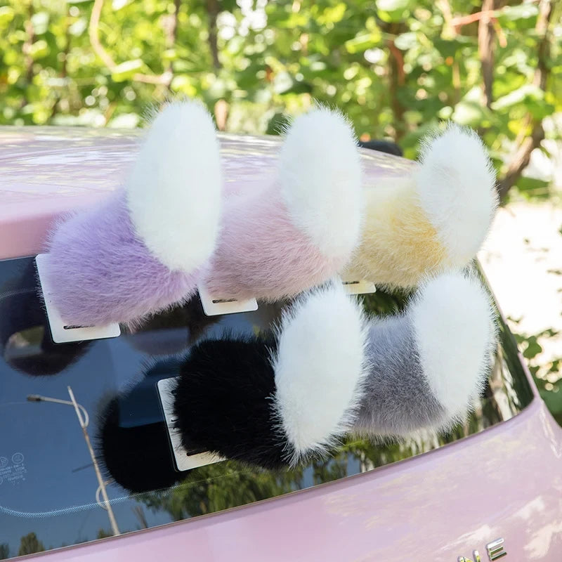 Five plush ear-like car moving tail accessories rest on a pink car's rear window, in purple, pink, yellow, black, and gray styles with white tips.