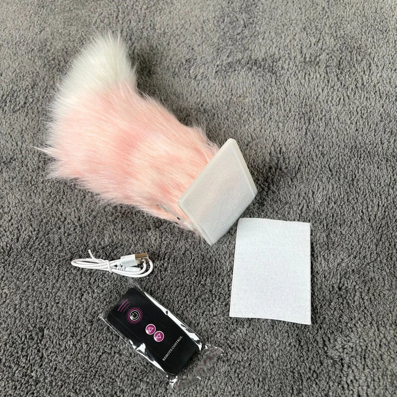 A pink and white plush car moving tail, a USB charging cable, a remote control, and an adhesive pad lie on a gray carpet.
