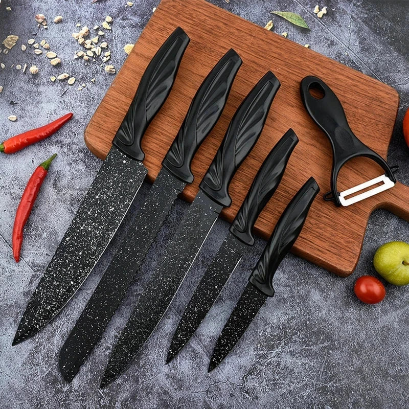 Five black speckled knives and a peeler from a kitchen utensil set laid on a wooden cutting board with chili peppers and fruit nearby.