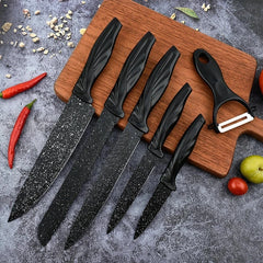 Five black speckled knives and a peeler from a kitchen utensil set laid on a wooden cutting board with chili peppers and fruit nearby.