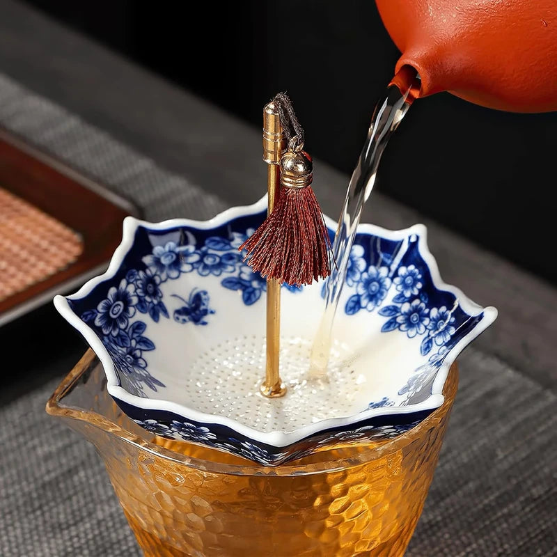 Red teapot pouring tea into a blue and white umbrella-shaped ceramic tea filter set over a textured glass pitcher
