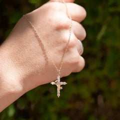 Faith Necklace - Gold crown cross pendant on fine chain held up against an out-of-focus green foliage background