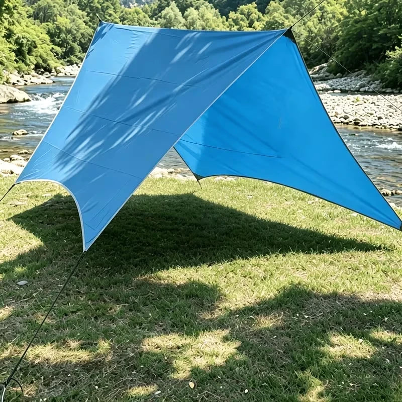 Mosquitero - Cobertizo de camping azul brillante instalado en una orilla herbosa del río, anclado con cuerdas, proyectando sombra sobre el césped.