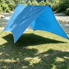 Mosquitero - Cobertizo de camping azul brillante instalado en una orilla herbosa del río, anclado con cuerdas, proyectando sombra sobre el césped.